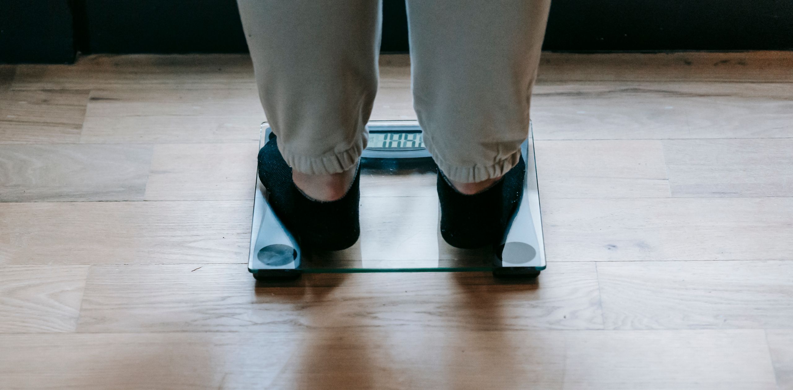 Back view of crop faceless obese female in socks weighing on glass scale placed on wooden floor in light room in daytime